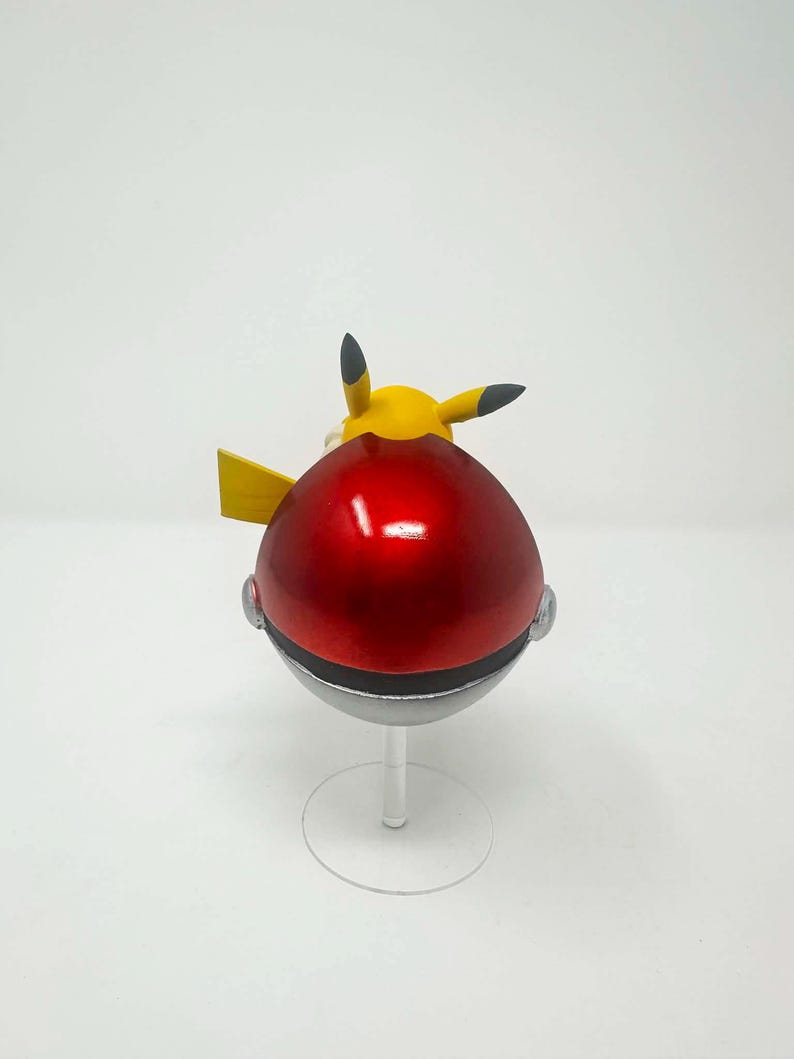 以下が含まれることがあります： 赤いと銀色のポケモンボールで、黄色いピカチュウのフィギュアが上にあります。ボールは光沢があり丸く、銀色のバンドと透明なスタンドが付いています。ピカチュウは黒い先端の耳と黄色の尾を持っています。