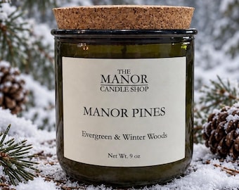 May include: A dark green glass candle jar with a cork lid. The white label reads: THE MANOR CANDLE SHOP, MANOR PINES, Evergreen & Winter Woods, Net Wt. 9 oz. The candle is positioned among pine branches and pinecones covered in fresh snow.