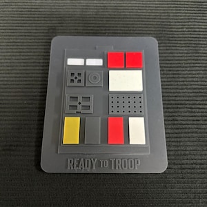 Op de afbeelding: Een grijs rechthoekig paneel met diverse gekleurde vormen en de tekst "READY TO TROOP". Het paneel heeft rode, witte, gele en grijze rechthoekige en vierkante vormen, samen met een reeks kleine cirkels.