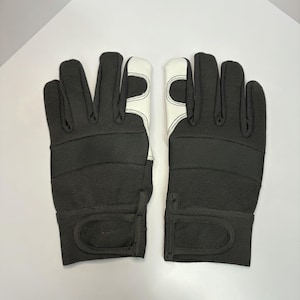Puede incluir: Un par de guantes negros y blancos. Los guantes tienen una tela negra acanalada en el dorso de la mano y los dedos, con material blanco en la palma. La muñeca tiene una correa negra con cierre de velcro.