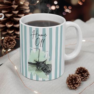 May include: White ceramic mug with vertical light blue stripes. The mug features the text "Home Off" and a watercolor illustration of a sleeping cat. The mug is filled with a dark liquid.