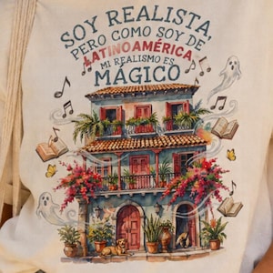 Può includere: Una t-shirt a maniche corte color bianco sporco con una grafica di un edificio a più piani con balconi, fiori e un cane. Il testo recita: SOY REALISTA, PERO COMO SOY DE LATINOAMÉRICA MI REALISMO ES MÁGICO, circondato da note musicali, libri e fantasmi.