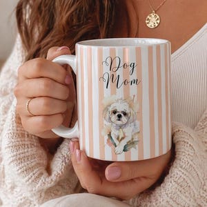 Può includere: Una tazza in ceramica bianca con strisce verticali color pesca e la scritta Dog Mom in corsivo nero. Sotto il testo si trova un'illustrazione in stile acquerello di un piccolo cane bianco e soffice con un accessorio al collo e fiori.