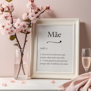 Può includere: Una stampa incorniciata bianca con la parola "Mãe" in nero, accompagnata da una definizione in portoghese. Un vaso di vetro con fiori rosa e un calice di champagne con liquido rosa sono presenti.