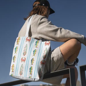 Può includere: Borsa tote bianca con un motivo a righe blu, con illustrazioni colorate di pesci e granchi. La borsa ha manici bianchi ed è realizzata in un tessuto resistente. Il design è ripetuto su tutta la superficie della borsa.