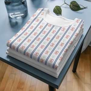 Può includere: Una pila di magliette bianche con un motivo a tema marino. La maglietta superiore presenta un motivo ripetuto di granchi rossi, strisce blu e alghe verdi su sfondo bianco. Le magliette sono ben piegate su un tavolo blu.