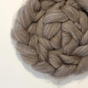 May include: Close-up of a circular, hand-braided wool rug in a neutral taupe color. The thick, soft wool is tightly woven, creating a textured surface. The rug's design features a repeating pattern of braided strands.