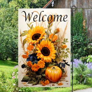Puede incluir: Una bandera de jardín de bienvenida con un tema otoñal. La bandera presenta girasoles, una calabaza y la palabra "Bienvenido" en una fuente de letra cursiva negra.