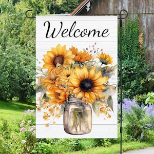 May include: A white wooden sign with the word "Welcome" in black script. A mason jar filled with a bouquet of sunflowers and greenery.