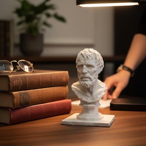 May include: A white bust of a man with a beard and short hair sits on a wooden desk next to a stack of books. The books are brown and red. A pair of spectacles rests on top of the books. A desk lamp is above the bust.