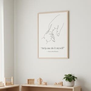 May include: A framed print featuring a minimalist line drawing of an adult's hand holding a child's hand. The print includes the quote "Help me do it myself" by Maria Montessori in a simple, elegant font. The frame is a light wood color.