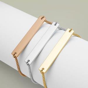 May include: Three bar bracelets in rose gold, silver, and gold. Each bracelet has a rectangular bar pendant attached to a fine chain. The bracelets are displayed on a white surface, highlighting their different metallic finishes.