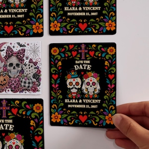 May include: Rectangular cards featuring black backgrounds with colorful floral borders, sugar skull illustrations, and the text ELARA & VINCENT NOVEMBER 21, 2027. One card displays a white background with a glittery skull, ghosts, and pumpkins.