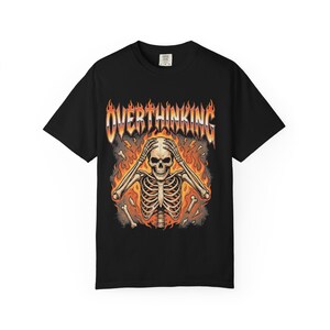 May include: A black short-sleeve t-shirt featuring a graphic print of a skeleton with hands on its head surrounded by orange and yellow flames. The word OVERTHINKING is printed in a metallic-style font above the skeleton, with scattered bone fragments.