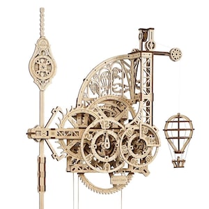 May include: A mechanical wooden wall clock model kit featuring intricate gear mechanisms, a pendulum, and a hanging hot air balloon weight. The clock face displays numbers and the text Aero Clock, with decorative hot air balloon cutouts integrated into the frame.