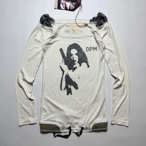 May include: An off-white long-sleeve shirt featuring a black graphic print of a person holding a firearm, with the text DPM printed nearby. The shirt has dark grey gathered shoulder details, a gold-toned interior label, and a hem with a drawstring.