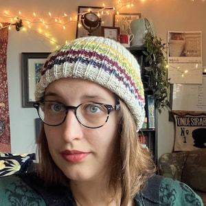 May include: A knit beanie featuring a ribbed texture and horizontal stripes in shades of cream, yellow, green, and dark blue. The hat has a folded brim and a multi-colored yarn construction, providing a snug fit around the head with a classic design.