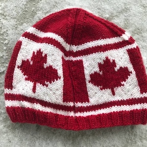 May include: A knitted beanie hat in red, white, and red. The hat displays the Canadian flag design, with a red maple leaf on a white background. The hat is made from a soft, knitted material.