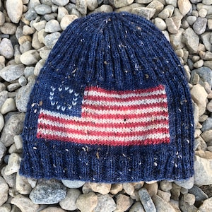 May include: A navy blue knit beanie featuring a vintage American flag design. The flag has red and white stripes and a field of white stars. The hat is speckled with flecks of white and brown.