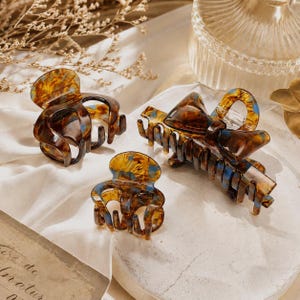 May include: A set of three hair clips featuring a tortoiseshell pattern with brown, amber, and blue marbled accents. The collection includes two small rounded claw clips and one larger rectangular claw clip adorned with a decorative bow detail.