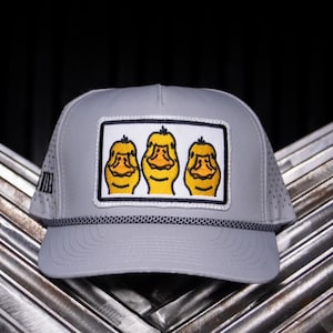 May include: A gray baseball cap with a perforated design and a black rope detail. The cap features a rectangular patch with three yellow cartoon faces. The background is dark.
