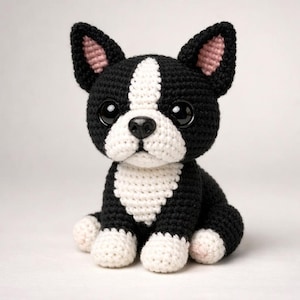 May include: A crocheted Boston Terrier dog with black and white markings, large black eyes, and pink inner ears. The handmade stuffed animal is sitting upright, showcasing its detailed craftsmanship and adorable design.