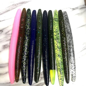 May include: A collection of ten soft plastic fishing lures arranged in a row. The lures are cylindrical with ribbed textures and come in various colors including solid pink, brown, dark green, blue, and translucent shades with glitter.