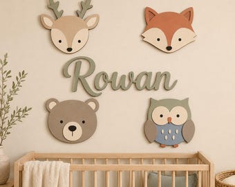 May include: A set of four wall-mounted animal cutouts featuring a deer, a fox, a bear, and an owl, arranged around the name Rowan written in a cursive green script. The animals are rendered in muted earth tones including beige, orange, and gray.