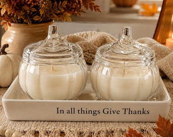 May include: Two clear glass pumpkin-shaped candle jars with lids, each containing white wax and a single wick. The jars sit on a rectangular white ceramic tray featuring the black printed text In all things Give Thanks on the front side.