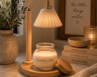 May include: A candle warmer lamp with a light wood base and stem, featuring a pleated fabric shade. A glass candle jar with the text Hearth and Lather sits on the base, accompanied by a round cork lid placed beside the jar.