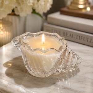 May include: A clear glass candle holder shaped like a seashell, featuring a scalloped rim and a spiral design. The container holds a white wax candle with a single lit wick, producing a warm, glowing flame that illuminates the textured glass surface.