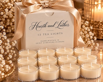 May include: A white square box labelled Hearth and Lather 12 Tea Lights Handcrafted in Saint Augustine Florida, topped with a gold satin ribbon. Twelve small round tea light candles in clear plastic cups are arranged in front of the box.