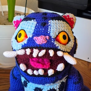 May include: A handmade blue crochet monster hat with large yellow and orange eyes, white teeth, and pink accents. The hat features a detailed design with a wide open mouth and two white tusks.