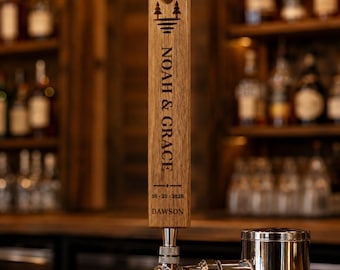 May include: A vertical wooden beer tap handle featuring a laser-engraved design with two pine trees, the text NOAH & GRACE, the date 06-20-2026, and the name DAWSON. The handle is mounted on a polished silver metal faucet base.