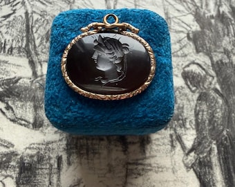 May include: An oval pendant with a dark cameo portrait set in a gold-toned frame. The pendant is displayed on a square of blue fabric. The background features a black and white sketch.