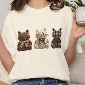 May include: A cream-coloured t-shirt featuring a graphic of three plush cats sitting in a row. The left cat has a brown leopard print, the middle cat is beige with brown polka dots, and the right cat has a black and white buffalo plaid pattern.