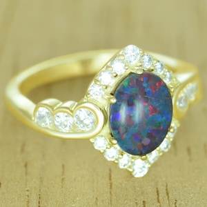 Op de afbeelding: Een gouden ring met een ovale, donkere opaal edelsteen met een levendig kleurenspel. De opaal is omgeven door een halo van kleine, heldere edelstenen. Extra heldere edelstenen zijn gezet langs de band van de ring.
