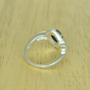 Op de afbeelding: Een zilveren ring met een donkere, ovaalvormige steen in het midden, omringd door kleinere, heldere stenen. De ringband heeft een decoratief ontwerp. De ring ligt op een lichtbruin houten oppervlak.