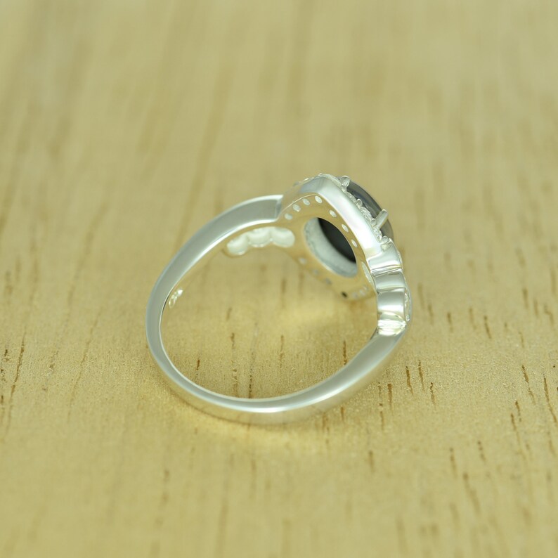 Op de afbeelding: Een zilveren ring met een donkere, ovaalvormige steen in het midden, omringd door kleinere, heldere stenen. De ringband heeft een decoratief ontwerp. De ring ligt op een lichtbruin houten oppervlak.