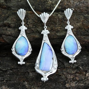 Puede incluir: Un conjunto de collar y pendientes de plata con piedras de ópalo azul iridiscente. Las joyas presentan diseños intrincados y ornamentados.