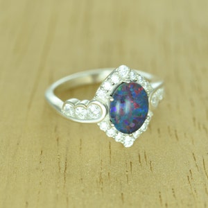 Op de afbeelding: Een zilveren ring met een ovale opaal edelsteen met een levendig kleurenspel. De opaal is omgeven door een halo van kleine, ronde, heldere edelstenen. Extra heldere edelstenen zijn gezet langs de band van de ring.