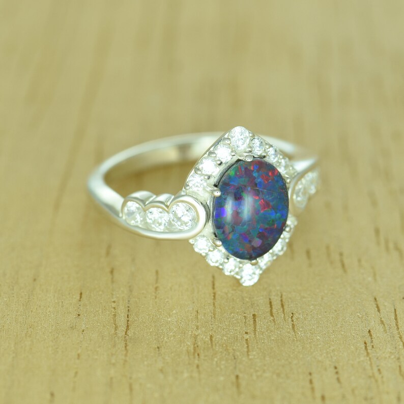 Op de afbeelding: Een zilveren ring met een ovale opaal edelsteen met een levendig kleurenspel. De opaal is omgeven door een halo van kleine, ronde, heldere edelstenen. Extra heldere edelstenen zijn gezet langs de band van de ring.