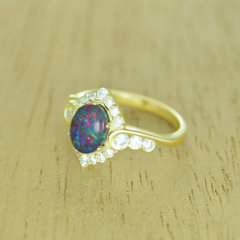 Op de afbeelding: Een gouden ring met een ovale, donkere opaal edelsteen met kleurflitsen. De opaal is omgeven door een halo van kleine, heldere edelstenen. De ringband is ook goud.