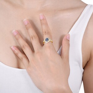 Op de afbeelding: Een gouden ring met een peervormige, blauw-paarse edelsteen in het midden, omringd door kleine, heldere stenen. De band van de ring is ook versierd met heldere stenen. De ring zit aan een vinger.
