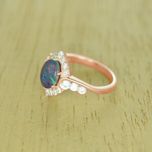 Op de afbeelding: Ros&eacute;gouden ring met een grote, ovale zwarte opaal met iriserende kleuren. De opaal is omgeven door kleine, heldere edelstenen. Verdere heldere edelstenen zijn langs de band van de ring gezet, wat een delicaat, elegant ontwerp cre&euml;ert. Een prachtig sieraad.