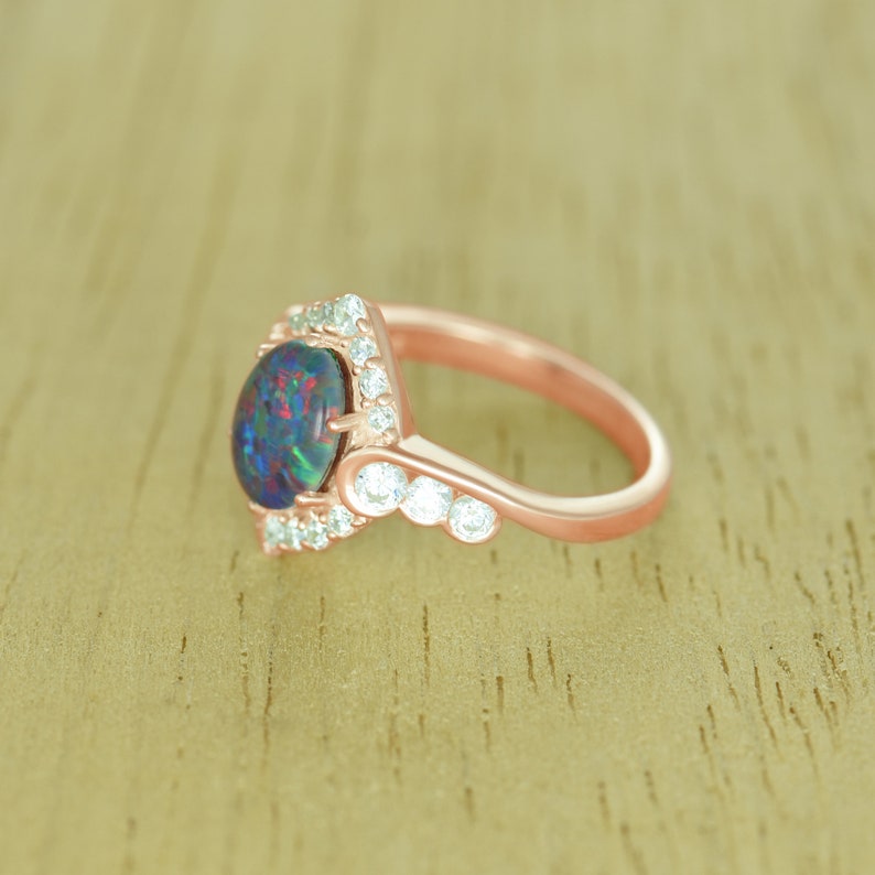 Op de afbeelding: Ros&eacute;gouden ring met een grote, ovale zwarte opaal met iriserende kleuren. De opaal is omgeven door kleine, heldere edelstenen. Verdere heldere edelstenen zijn langs de band van de ring gezet, wat een delicaat, elegant ontwerp cre&euml;ert. Een prachtig sieraad.