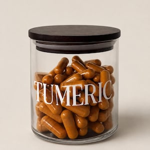 May include: A clear glass jar with a dark wooden lid, filled with orange turmeric capsules. The word "TURMERIC" is printed in white on the front of the jar. The jar is filled with capsules.