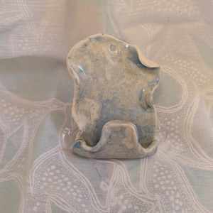 Può includere: Un porta telefono in ceramica fatto a mano nei toni del blu e del bianco. Il supporto ha una forma organica unica con un piccolo foro nella parte superiore. La superficie ha un aspetto maculato e strutturato. La base ha un piccolo bordo per tenere un telefono.