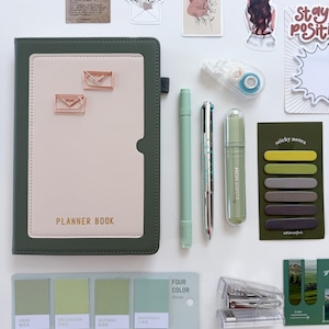 May include: An assortment of stationery items on a white surface. Includes a green and beige planner book, pens, sticky notes, a stapler, and stickers. The planner book reads "PLANNER BOOK". The color palette is primarily green and beige.