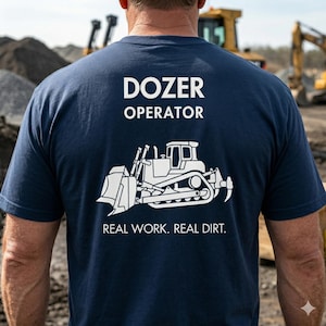 Könnte beinhalten: Marineblaues T-Shirt mit weißem Text und einer Bulldozer-Grafik. Der Text lautet "DOZER OPERATOR" über der Grafik und "REAL WORK. REAL DIRT." darunter. Das Shirt besteht aus einem weichen Material.